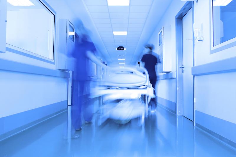 Eine blaue Aufnahme eines Krankenhausganges in welchem zwei Pflegepersonen ein Krankenbett schieben.
